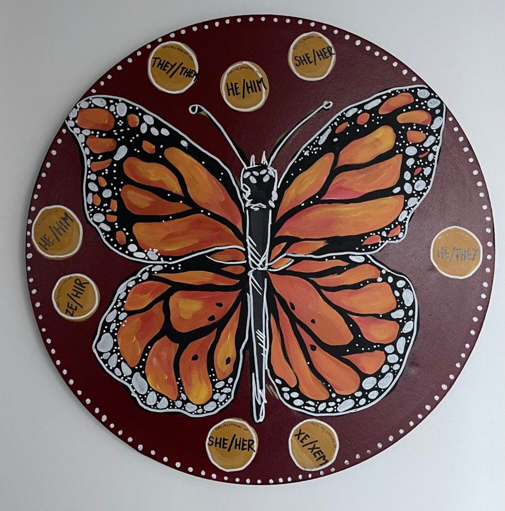 A brown disc with a monarch butterfly, centred, painted on it. Surrounding the monarch butterfly are smaller painted discs with pronouns in them. In clockwise order, these are: they/them, he/him, sh/her, he/they, xe/xem, she/her.
