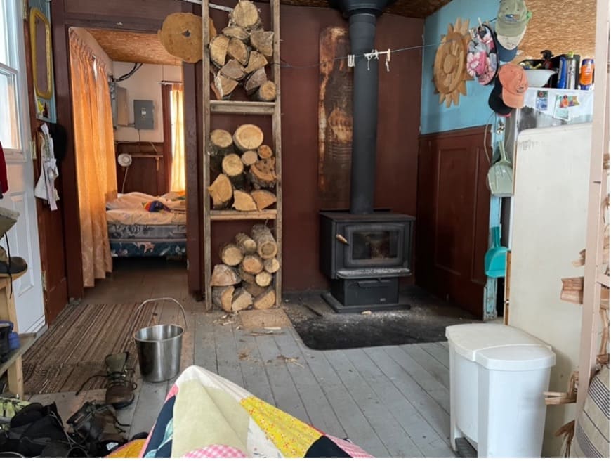 The view of the inside of Claude's cottage from his chair. Through an open doorway, part of a bed can be seen. In the same room as the photographer, a pile of firewood can be seen next to a wood stove.